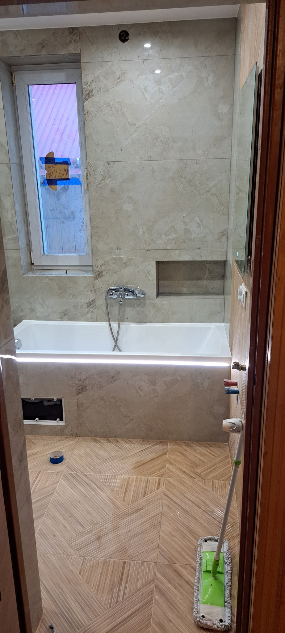 Wykończona łazienka z wanną w beżowych płytkach, oświetleniem LED i wzorzystą podłogą. Widok zza uchylonych drzwi, mop na podłodze.