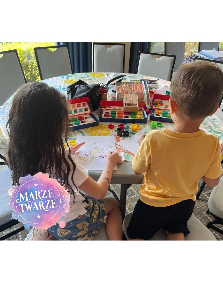Dwoje dzieci maluje farbami obrazki na papierze przy okrągłym stole, na którym znajdują się zestawy farb i pędzle.