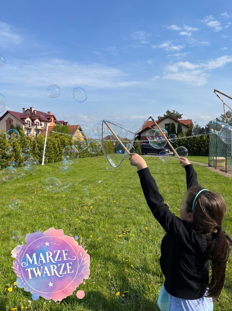 Dziewczynka w czarnej bluzie puszcza bańki mydlane za pomocą dwóch kijów z siatką na trawniku w słoneczny dzień, w tle domy i zielone drzewa.