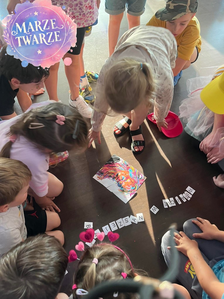 Grupa dzieci siedzi na podłodze wokół rozłożonej układanki z obrazkiem lwa i liter układających się w napis, prawdopodobnie podczas zabawy lub warsztatów.