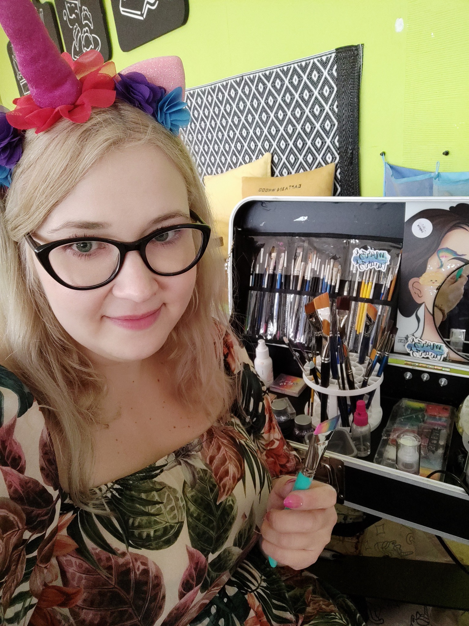 Uśmiechnięta blondynka w okularach i opasce jednorożca, trzymająca pędzel do makijażu, obok kufer z akcesoriami do malowania twarzy.