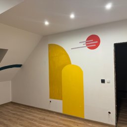 olimp-bud paweł czaja - Wnętrze pokoju na poddaszu z drewnianą podłogą i pomalowanymi ścianami w geometryczne wzory: żółte kształty i czerwone koło z szarymi liniami. Widoczne gniazdka elektryczne i otwór drzwiowy bez...