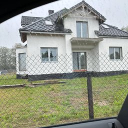olimp-bud paweł czaja - Niedokończony biały dom murowany z szarą dachówką, widziany przez ogrodzenie z siatki, z perspektywy wnętrza samochodu w pochmurny dzień.