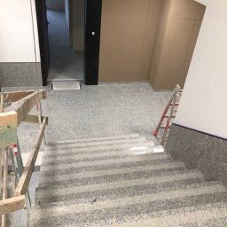 olimp-bud paweł czaja - Klatka schodowa w trakcie remontu: widoczne zabezpieczone schody, drabina oparta o ścianę oraz otwarte drzwi do dalszej części budynku.