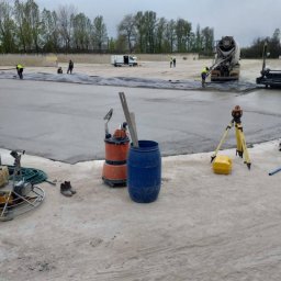 olimp-bud paweł czaja - Wylewka betonowa na dużej powierzchni, widoczny mikser do betonu, pracownicy, poziomica laserowa, pompa do wody i narzędzia budowlane.