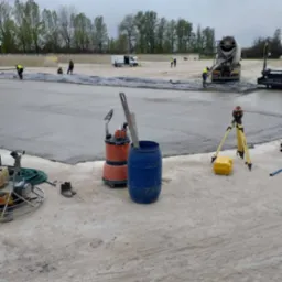 Wylewka betonowa na dużej powierzchni, widoczny mikser do betonu, pracownicy, poziomica laserowa, pompa do wody i narzędzia budowlane.