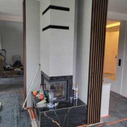 olimp-bud paweł czaja - Wnętrze w trakcie wykańczania z nowoczesnym kominkiem z czarnym marmurem i betonową obudową z poziomymi wnękami, widoczne materiały budowlane i folia ochronna na podłodze.