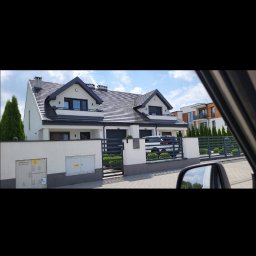 olimp-bud paweł czaja - Nowoczesny dom jednorodzinny z ciemnym dachem i jasną elewacją, widoczny zza ogrodzenia z metalowymi przęsłami i betonowymi słupkami, z zielonymi tujami posadzonymi wzdłuż ogrodzenia.