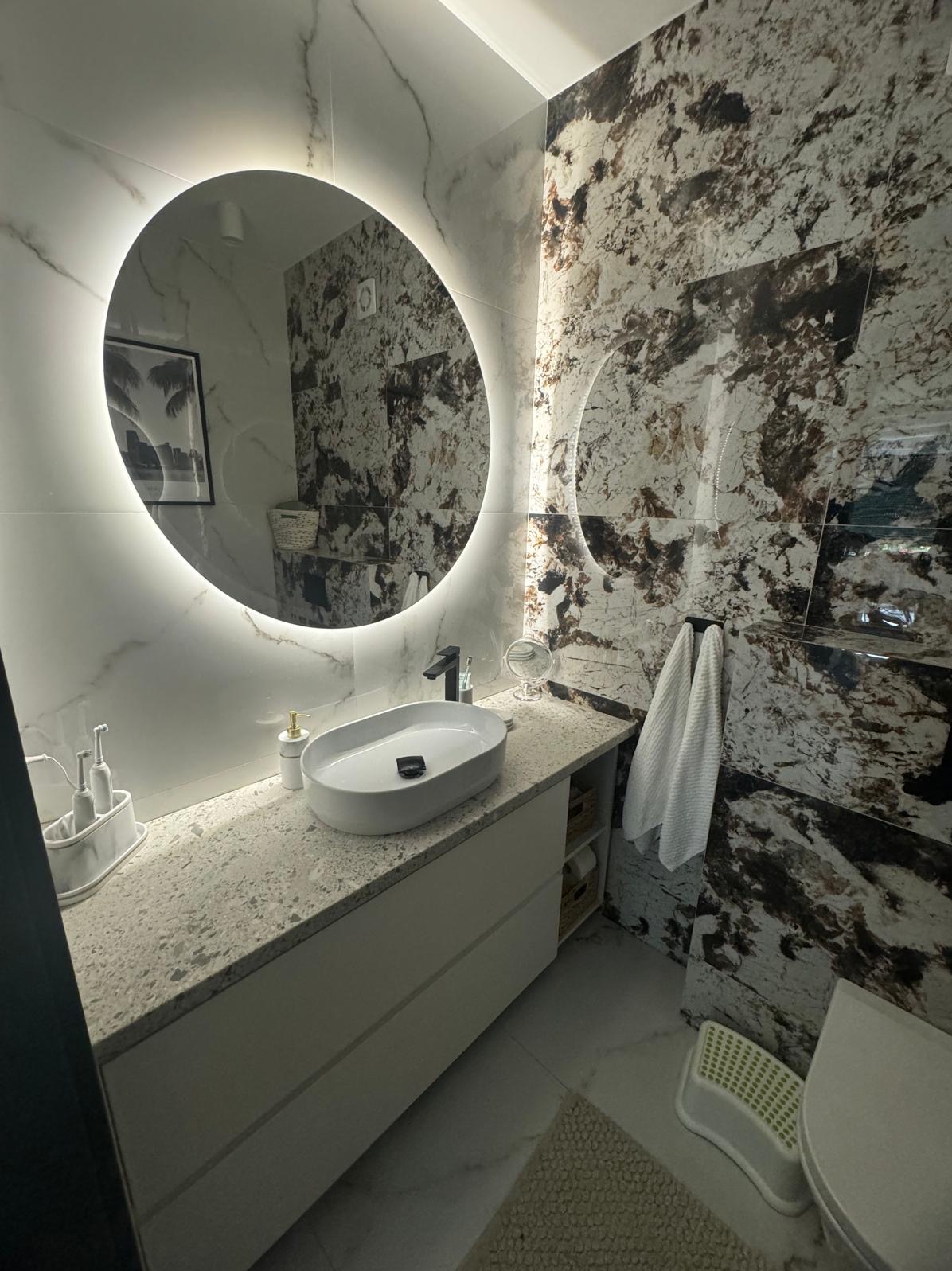 Nowoczesna łazienka z owalną umywalką nablatową, podświetlanym okrągłym lustrem, marmurowymi płytkami na ścianach i białymi meblami pod umywalką.