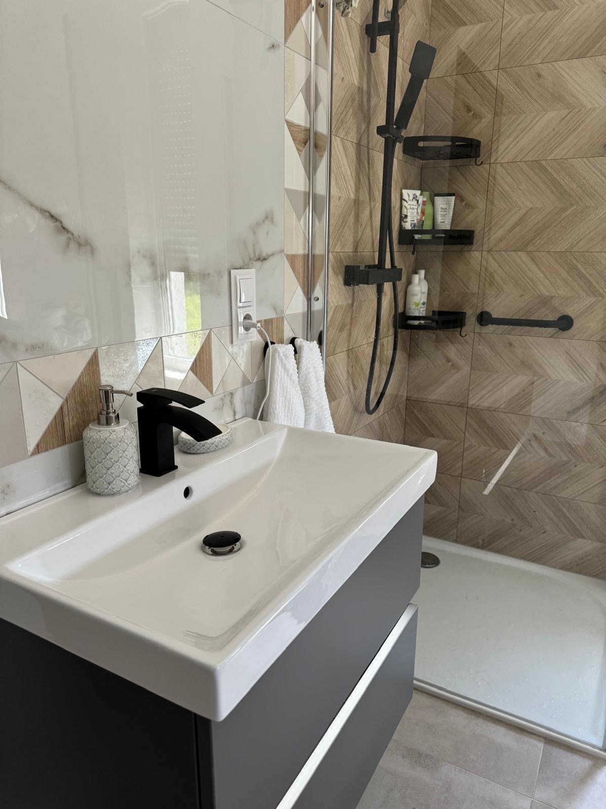 Nowoczesna łazienka z umywalką nablatową, czarną armaturą i kabiną prysznicową z drewnopodobnymi płytkami w jodełkę.