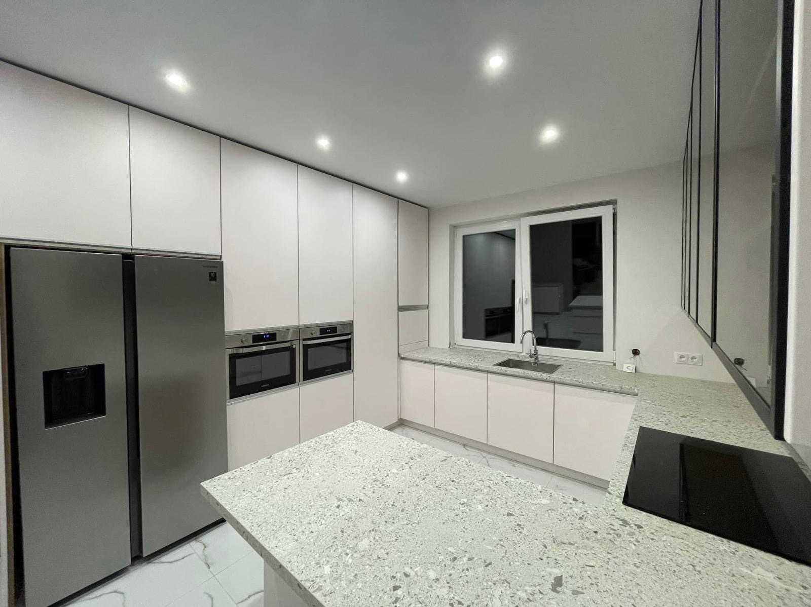 Nowoczesna kuchnia z białymi szafkami, szarym blatem z konglomeratu, zabudowanym sprzętem AGD (lodówka side-by-side, piekarniki) i wyspą kuchenną z płytą indukcyjną.