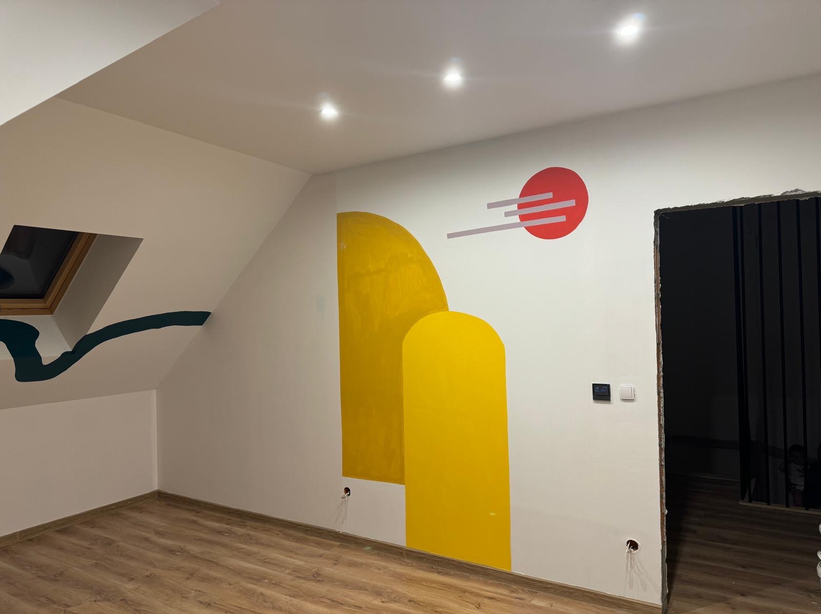 Wnętrze pokoju na poddaszu z drewnianą podłogą i pomalowanymi ścianami w geometryczne wzory: żółte kształty i czerwone koło z szarymi liniami. Widoczne gniazdka elektryczne i otwór drzwiowy bez...
