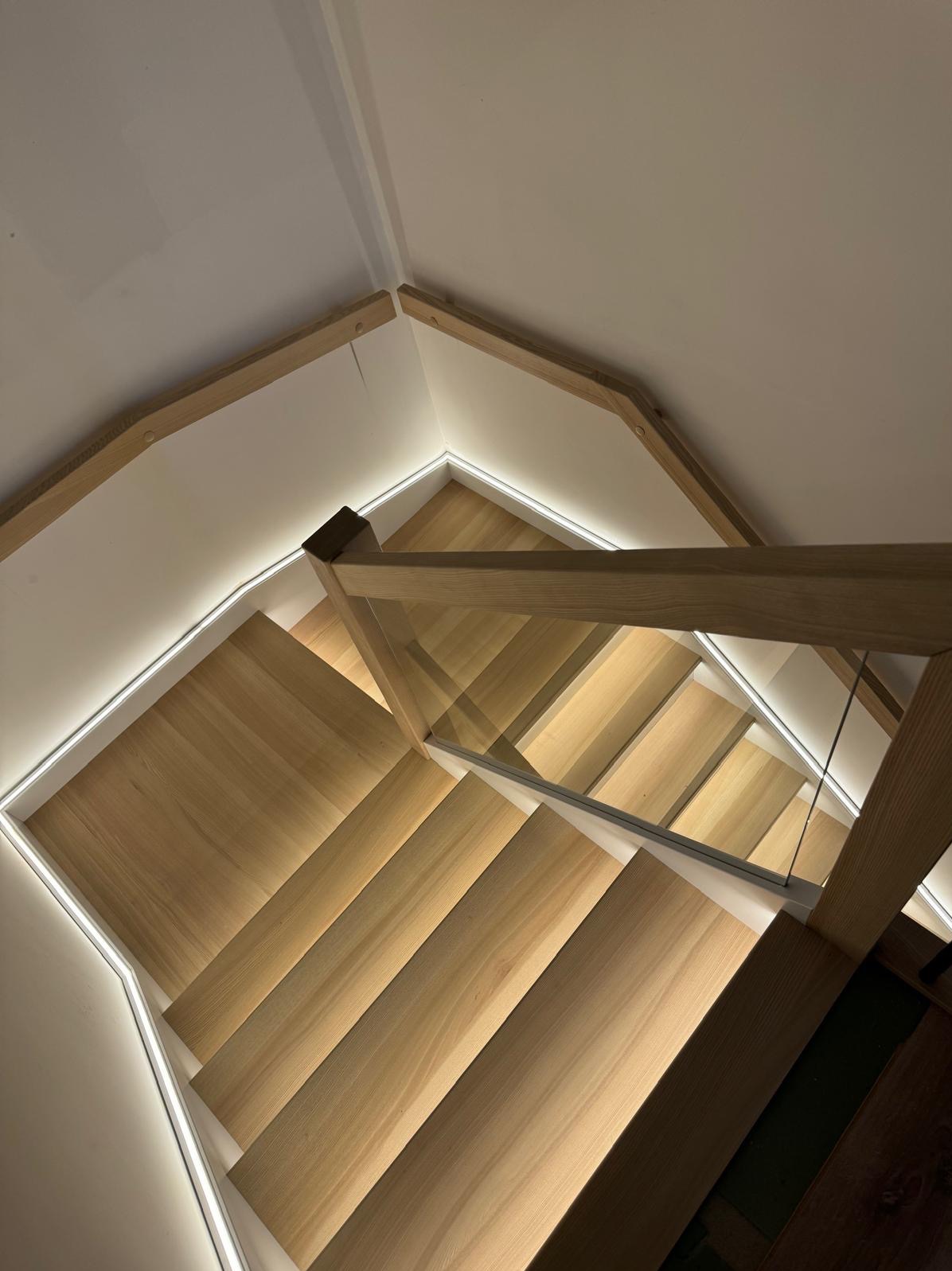 Widok z góry na drewniane schody z podświetleniem LED i szklaną balustradą, z widocznym fragmentem podłogi na dole.