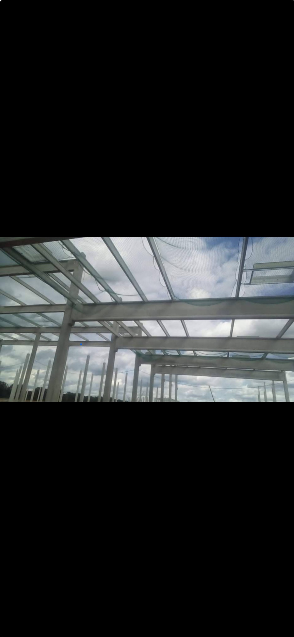 Konstrukcja szkieletowa budynku w trakcie budowy, widoczne stalowe belki i słupy, siatka zabezpieczająca oraz niebo w tle.