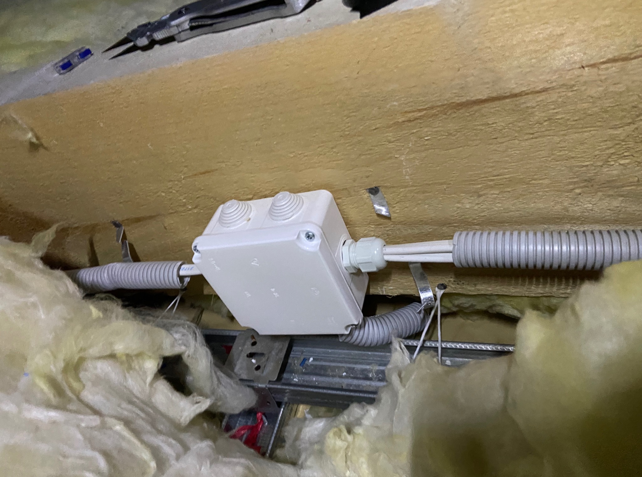 Biała puszka elektryczna z szarymi peszlami przymocowana do drewnianej konstrukcji, otoczona izolacją, narzędzia w tle. Instalacja w trakcie prac.