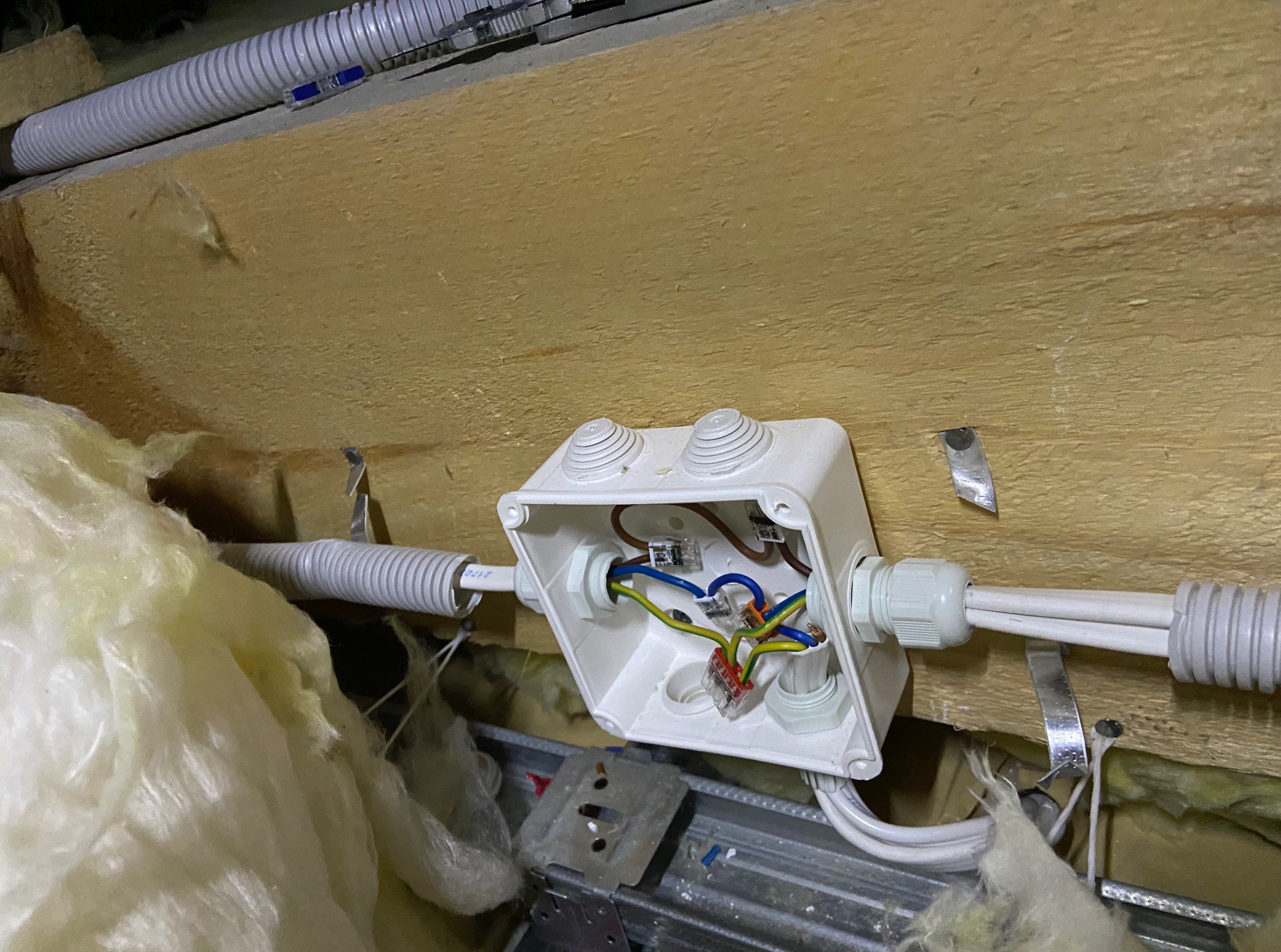 Otwarta, biała puszka elektryczna z widocznymi przewodami w kolorach: brązowym, niebieskim, zielono-żółtym. Kable wchodzą do puszki przez elastyczne peszle.