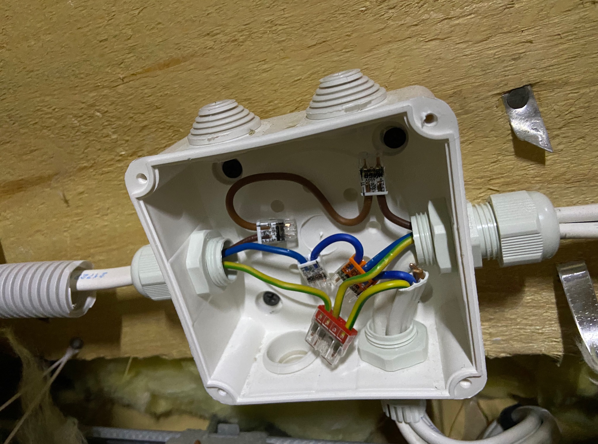 Biała puszka elektryczna z widocznymi przewodami i złączkami, zamontowana na drewnianej powierzchni. Detal instalacji w trakcie realizacji.