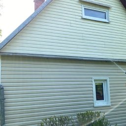 ECOMMTECH JAGIENKA JECZEŃ-PORADA - Czyszczenie sidingu domu z użyciem myjki ciśnieniowej. Woda spływa po elewacji, widoczne okna i fragment zieleni. Jasne, naturalne oświetlenie.