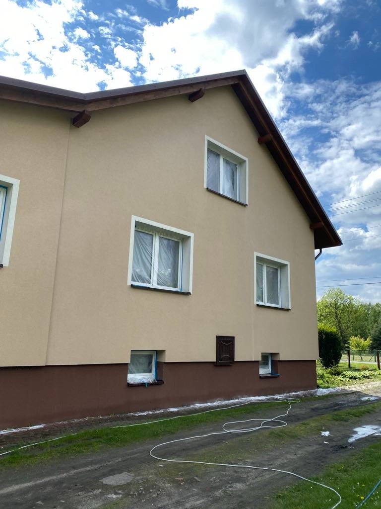 Dwukondygnacyjny dom z beżową elewacją, okna zabezpieczone folią, widoczny wąż do mycia. Przygotowania do mycia elewacji pod kątem.
