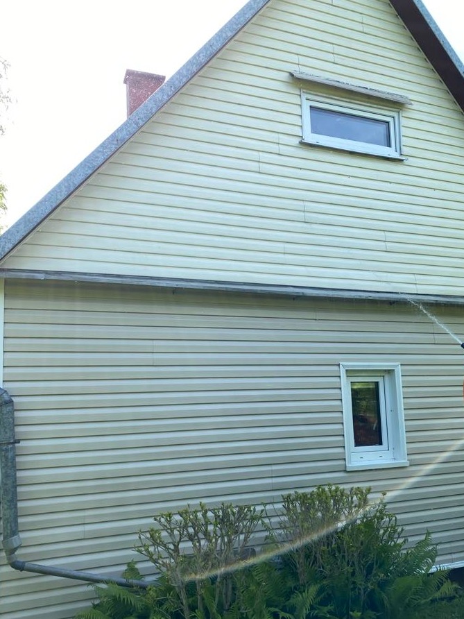 Czyszczenie sidingu domu z użyciem myjki ciśnieniowej. Woda spływa po elewacji, widoczne okna i fragment zieleni. Jasne, naturalne oświetlenie.