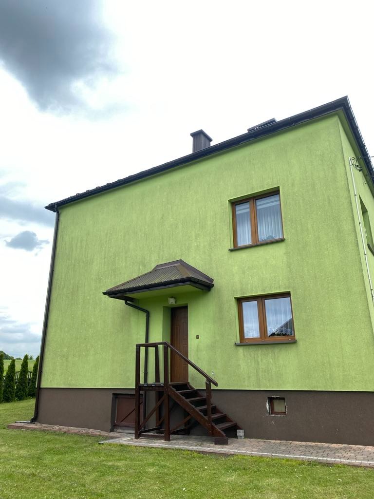 Dwupiętrowy dom z zieloną elewacją i brązowymi oknami w Rzeszowie, widziany z dołu. Schody prowadzą do wejścia, a niebo jest pochmurne.