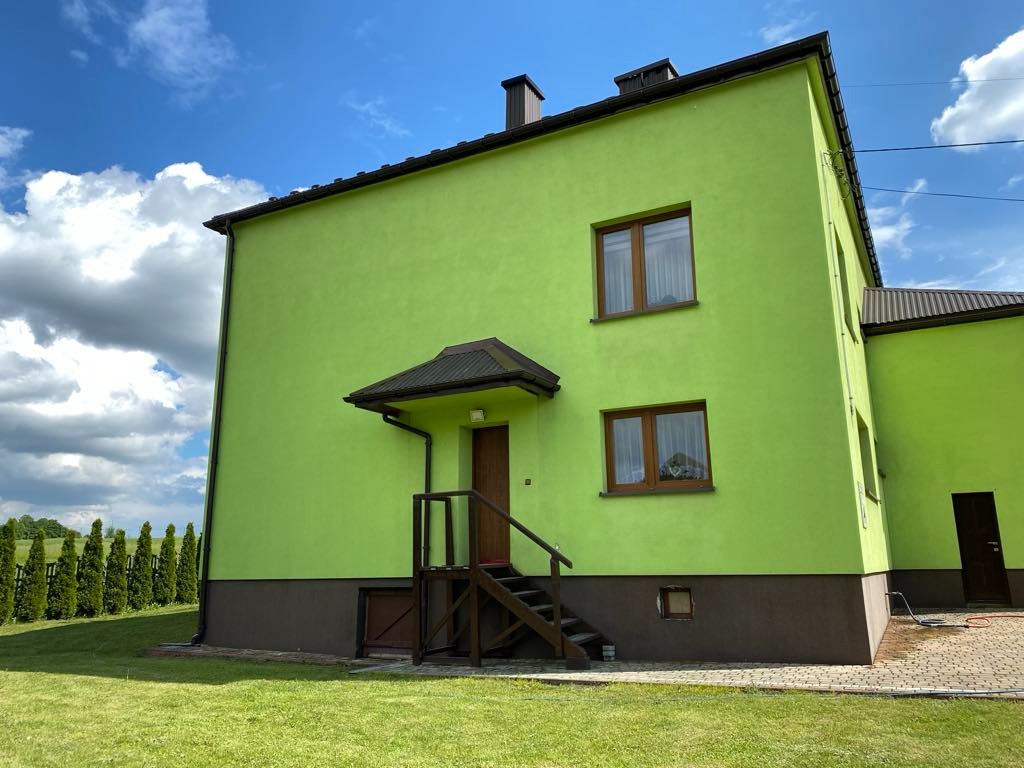 Dwukondygnacyjny dom z zieloną elewacją i brązowymi akcentami, widoczny z perspektywy ogrodu. Uwagę przyciągają drewniane schody i okna.