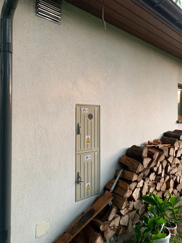 Elewacja budynku z szarą rynną, skrzynką ZPL-1/ZK-1 i stosem drewna opartym o ścianę. Widoczny fragment okna i wentylacja.