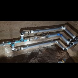 Firma Usługowa Łukasz Głowacki - Instalacja wodociągowa z szarych rur PVC, z licznymi złączkami i zaworami, zamontowana w betonowym wykopie, widoczne ślady ziemi.