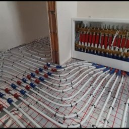 Firma Usługowa Łukasz Głowacki - Rozłożone białe rury ogrzewania podłogowego na siatce, z czerwono-niebieskimi złączkami, prowadzące do rozdzielacza w białej obudowie ściennej.