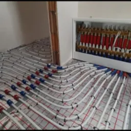 Rozłożone białe rury ogrzewania podłogowego na siatce, z czerwono-niebieskimi złączkami, prowadzące do rozdzielacza w białej obudowie ściennej.