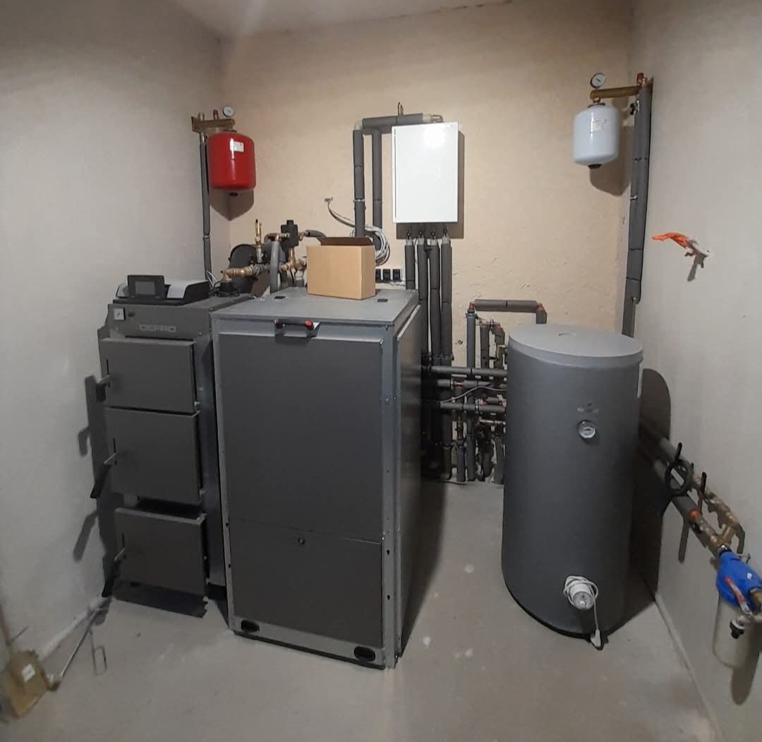 Kompletna kotłownia z piecem na paliwo stałe, zasobnikiem ciepłej wody, naczyniami wzbiorczymi i osprzętem hydraulicznym, w pomieszczeniu o surowych ścianach i betonowej podłodze.