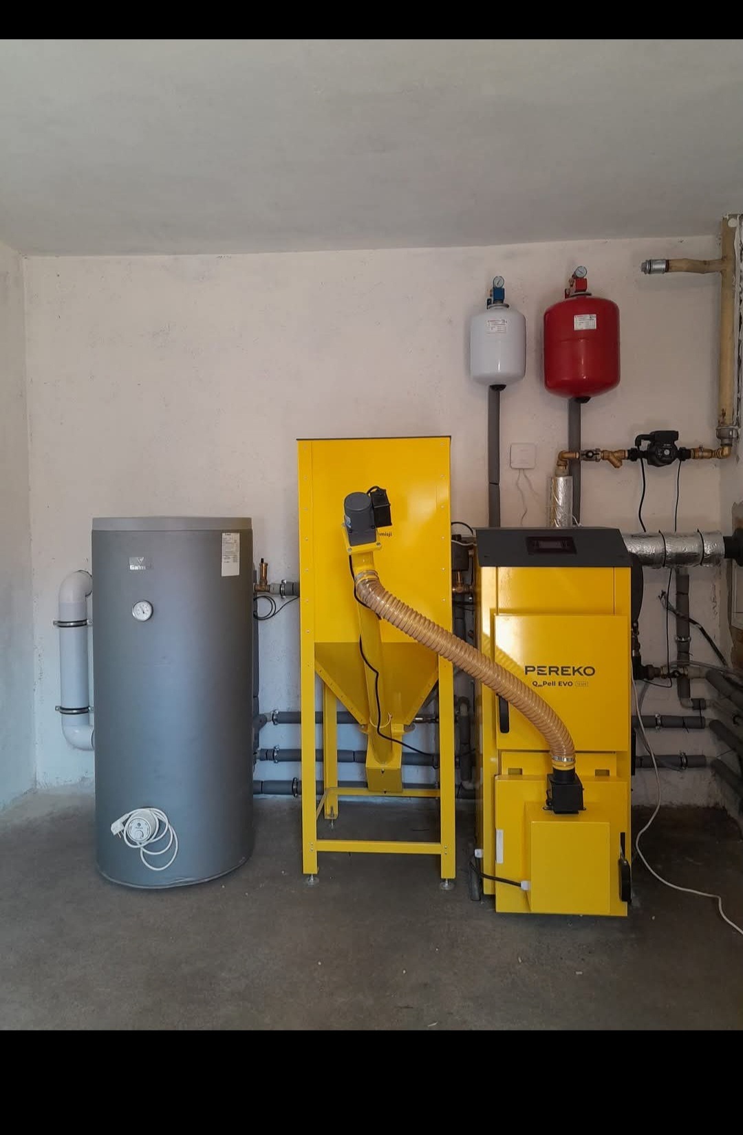 Szary zasobnik ciepłej wody użytkowej obok żółtego podajnika i kotła na pellet Pereko w kotłowni z białymi ścianami i instalacjami grzewczymi.