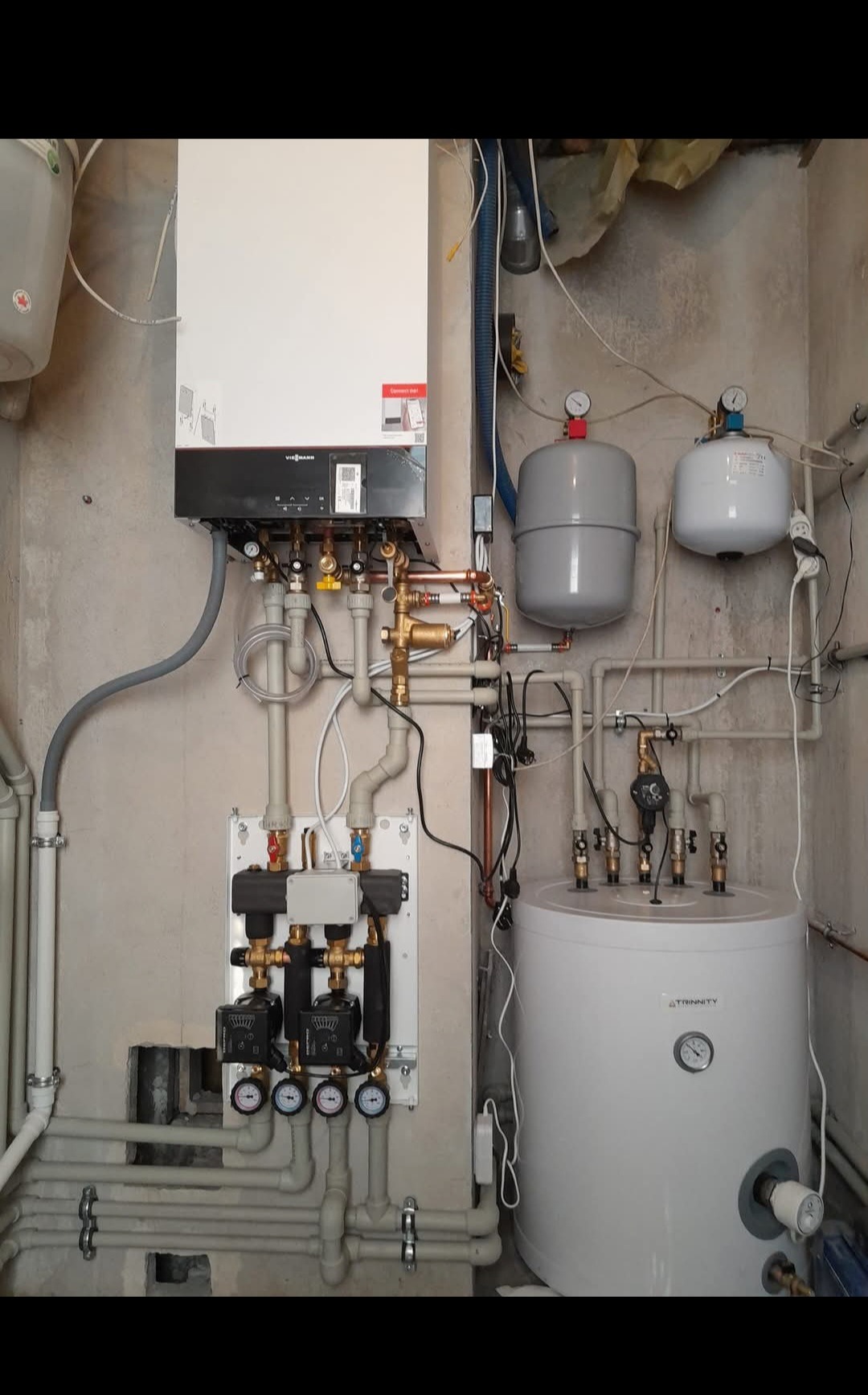 Nowoczesny piec CO Viessmann z osprzętem, w tym biały zasobnik ciepłej wody Trinity i dwa zbiorniki wyrównawcze, zamontowany na surowej, betonowej ścianie w kotłowni.