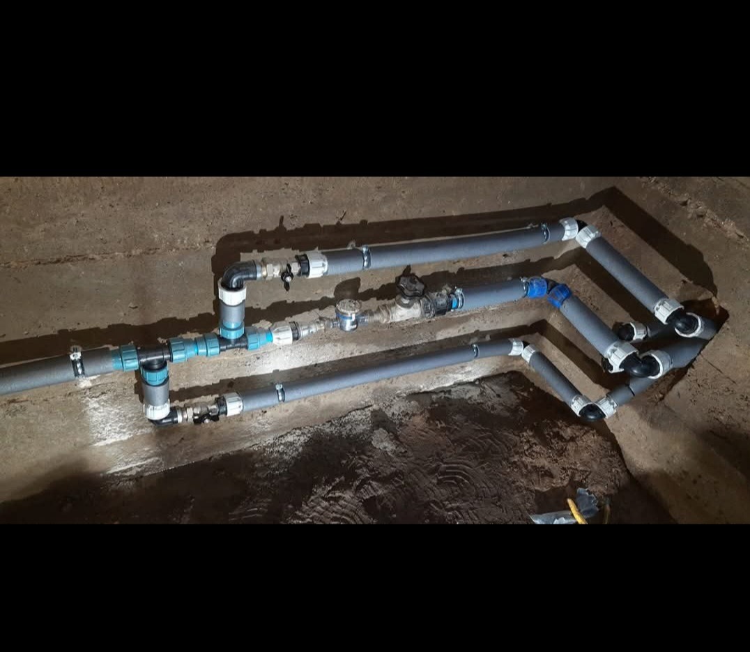 Instalacja wodociągowa z szarych rur PVC, z licznymi złączkami i zaworami, zamontowana w betonowym wykopie, widoczne ślady ziemi.