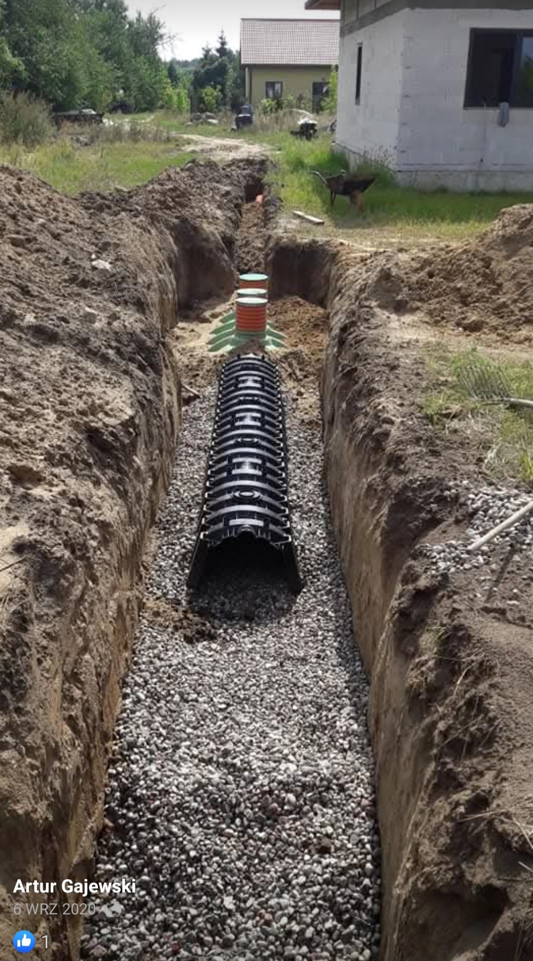 Wykop z ułożonym drenażem rurowym obsypanym żwirem, widoczne studzienki kontrolne i otoczenie działki z budynkiem.
