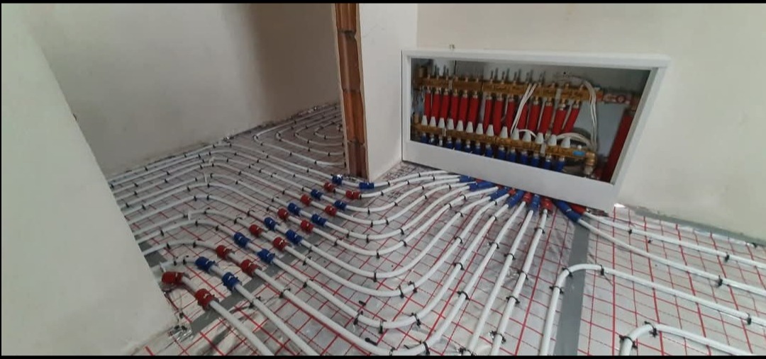 Rozłożone białe rury ogrzewania podłogowego na siatce, z czerwono-niebieskimi złączkami, prowadzące do rozdzielacza w białej obudowie ściennej.