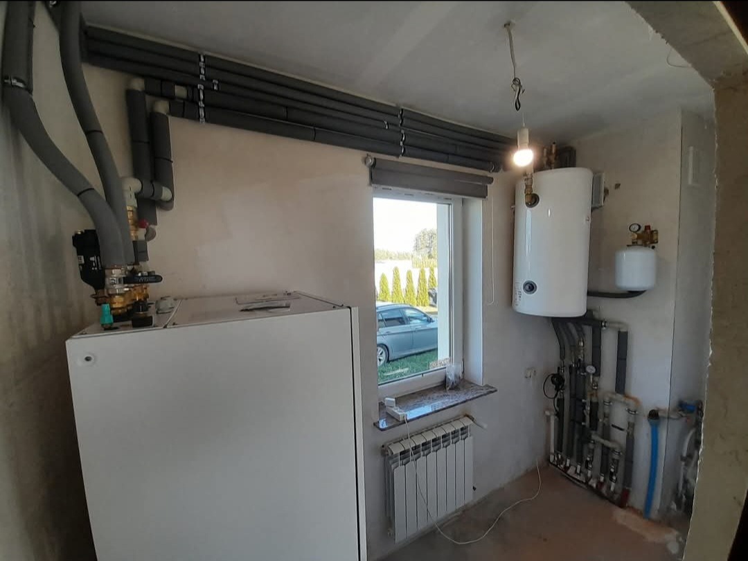 Kotłownia w trakcie montażu, widoczne rury grzewcze z izolacją, zasobnik ciepłej wody i grzejnik, surowy stan ścian.