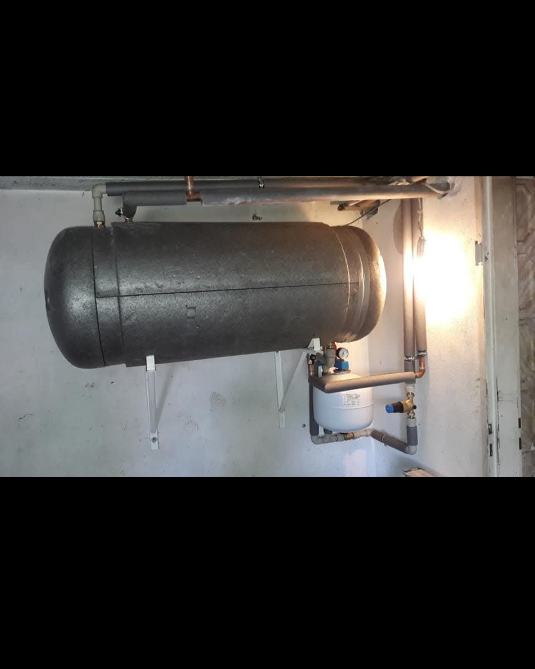 Poziomy, zaizolowany zbiornik akumulacyjny ciepła zamontowany na ścianie, z widocznym naczyniem wzbiorczym i rurami w izolacji, oświetlony z boku.