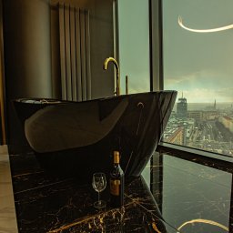 AR interior design- projektowanie wnętrz Angelika Rucka - Luksusowa czarna wanna z złotą armaturą na tle panoramy miasta z wysokiego piętra. Butelka wina i kieliszek dodają elegancji.