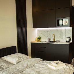 AR interior design- projektowanie wnętrz Angelika Rucka - Czarne szafki kuchenne z marmurowym blatem i złotą armaturą, widoczne z perspektywy łóżka w pokoju hotelowym. Minimalistyczny design, oświetlenie LED.