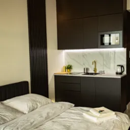 Czarne szafki kuchenne z marmurowym blatem i złotą armaturą, widoczne z perspektywy łóżka w pokoju hotelowym. Minimalistyczny design, oświetlenie LED.