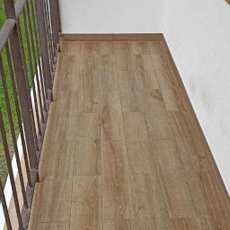 Marek - Wykończenia wnętrz, usługi remontowo budowlane - Warszawa i okolice - Wykończony balkon z panelami podłogowymi imitującymi drewno, widoczne metalowe barierki i fragment białej ściany.