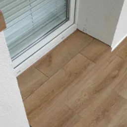 Ułożone panele podłogowe w odcieniach dębu, widoczne przy białym oknie z żaluzjami i fragmentem ściany z nier&oacute;wną fakturą.