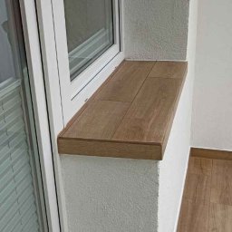 Marek - Wykończenia wnętrz, usługi remontowo budowlane - Warszawa i okolice - Wykończony parapet z płytek imitujących drewno na balkonie w Jabłonnie, dopasowany do podłogi z paneli. Widoczna biała rama okienna i struktura ściany.