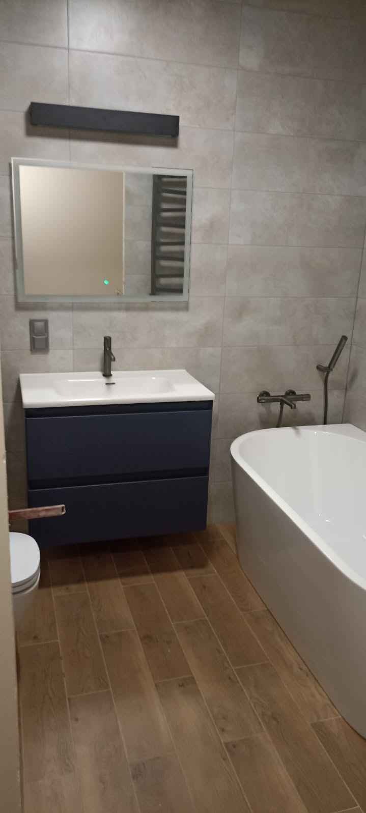 Nowoczesna łazienka z drewnopodobną podłogą, wiszącą szafką umywalkową w kolorze granatowym, wanną wolnostojącą i szarą glazurą ścienną.