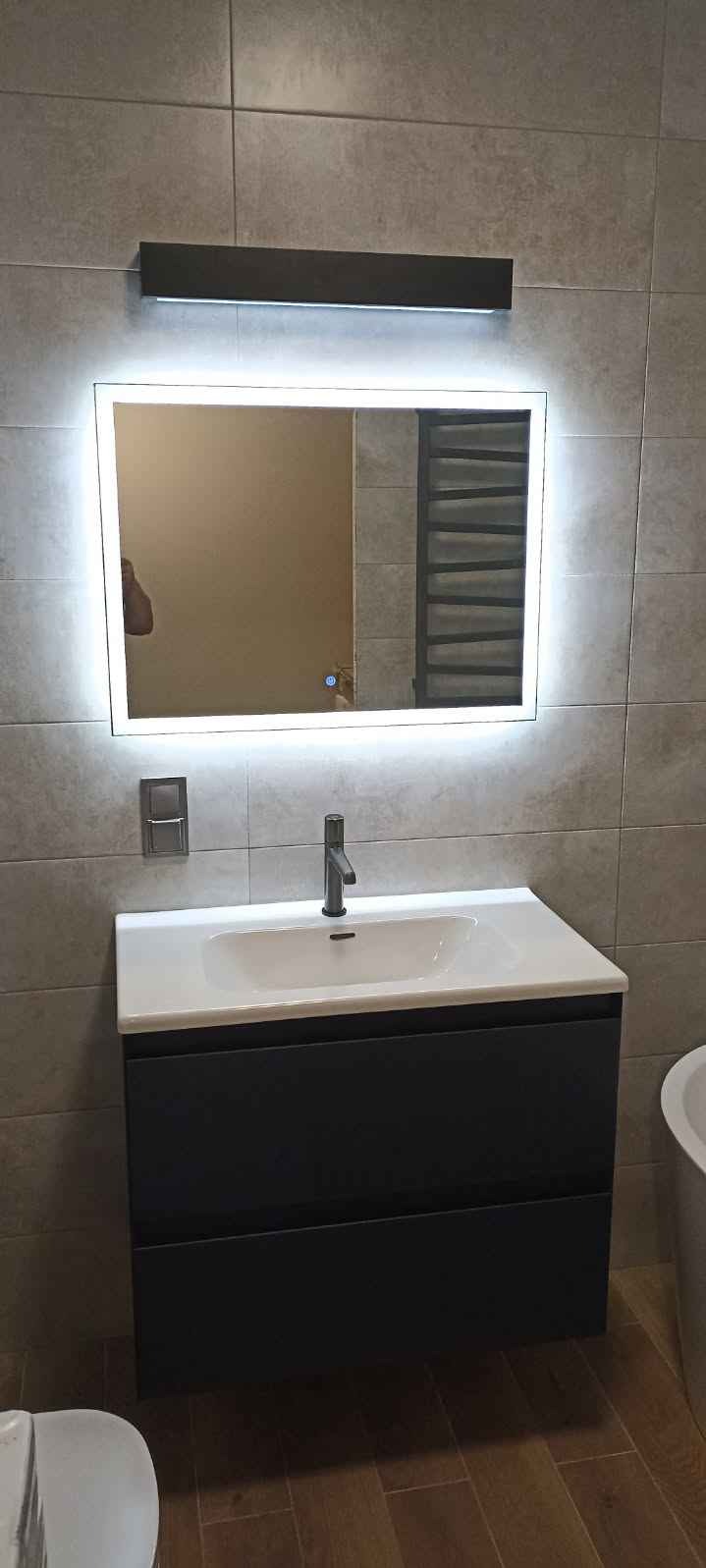 Nowoczesna umywalka z szafką w kolorze antracytowym, podświetlane lustro LED na szarej ścianie z płytek, bateria w kolorze grafitowym, drewnopodobna podłoga