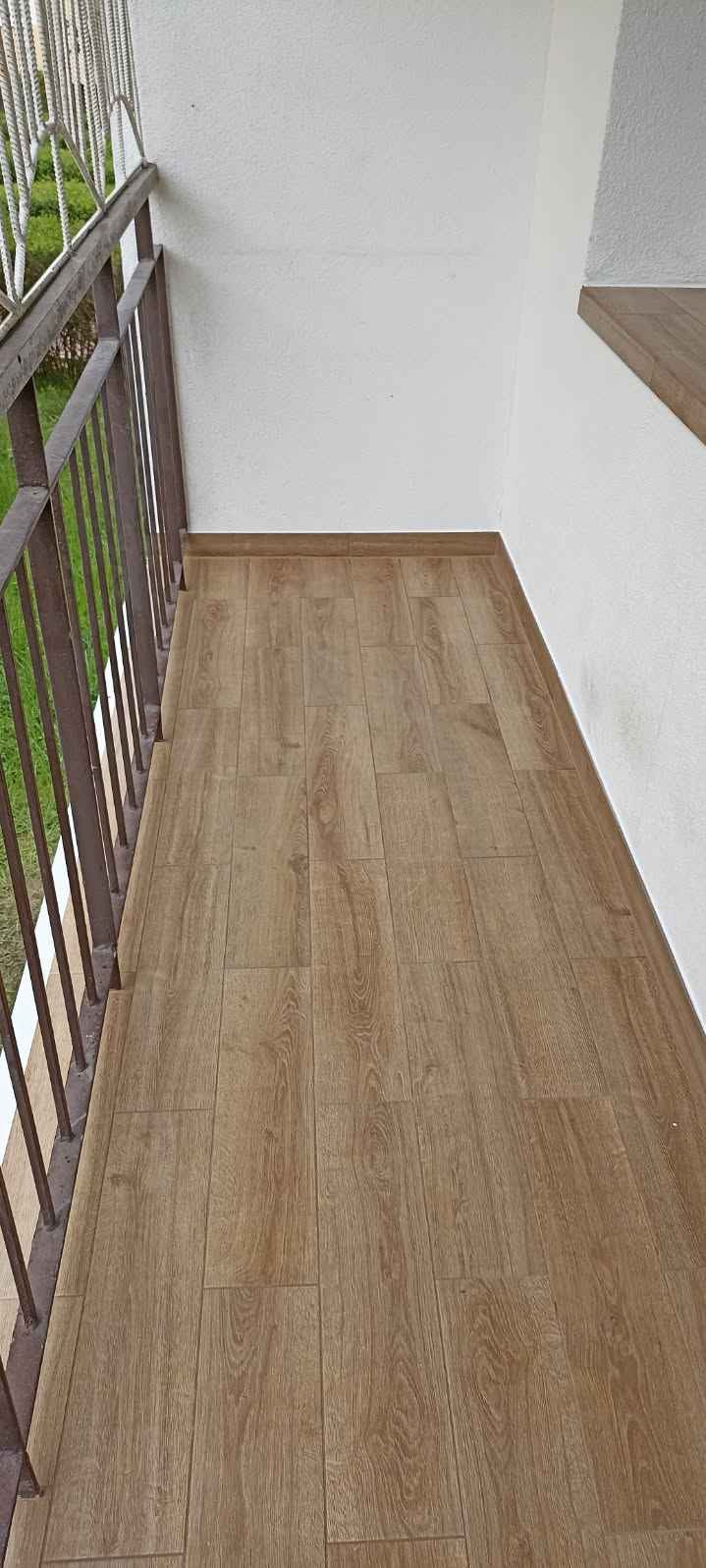 Wykończony balkon z panelami podłogowymi imitującymi drewno, widoczne metalowe barierki i fragment białej ściany.