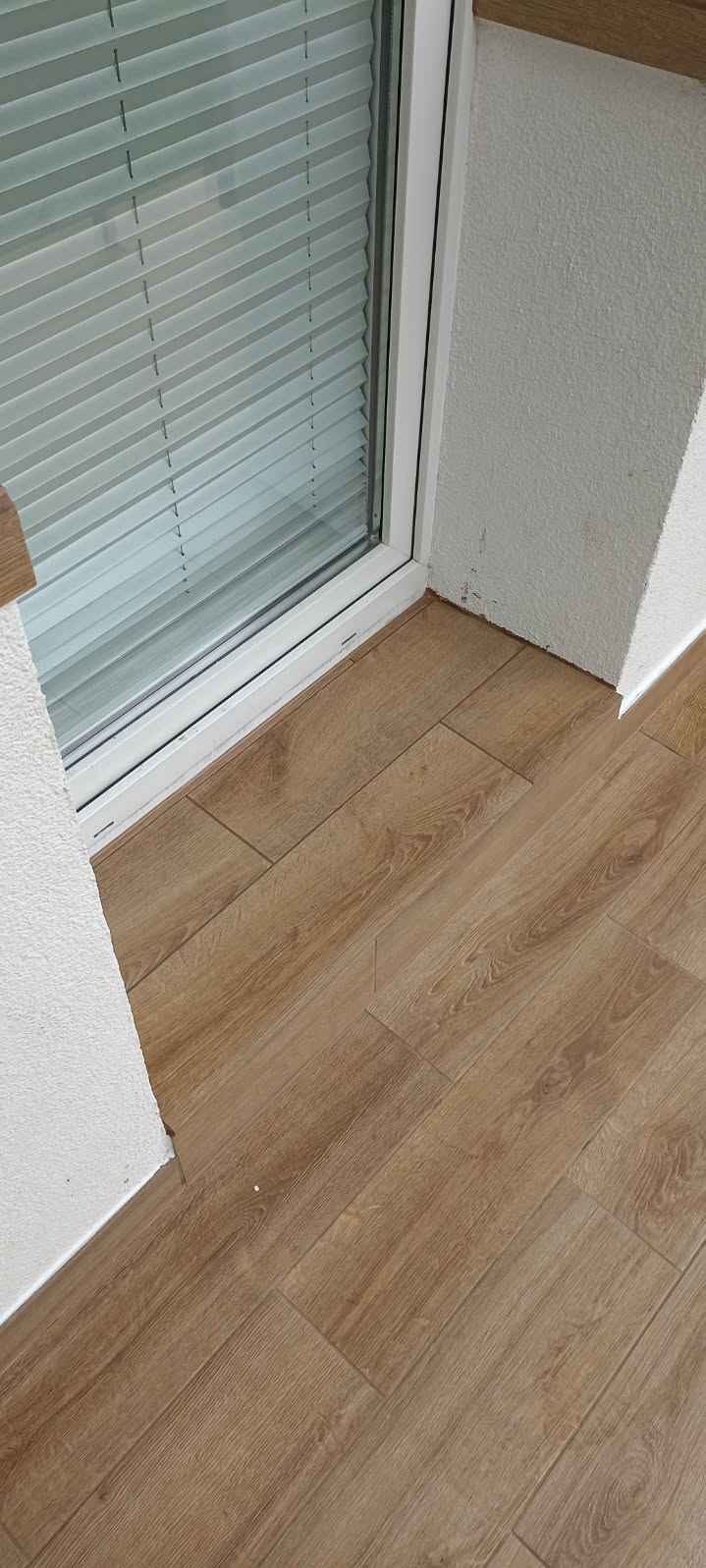 Ułożone panele podłogowe w odcieniach dębu, widoczne przy białym oknie z żaluzjami i fragmentem ściany z nierówną fakturą.