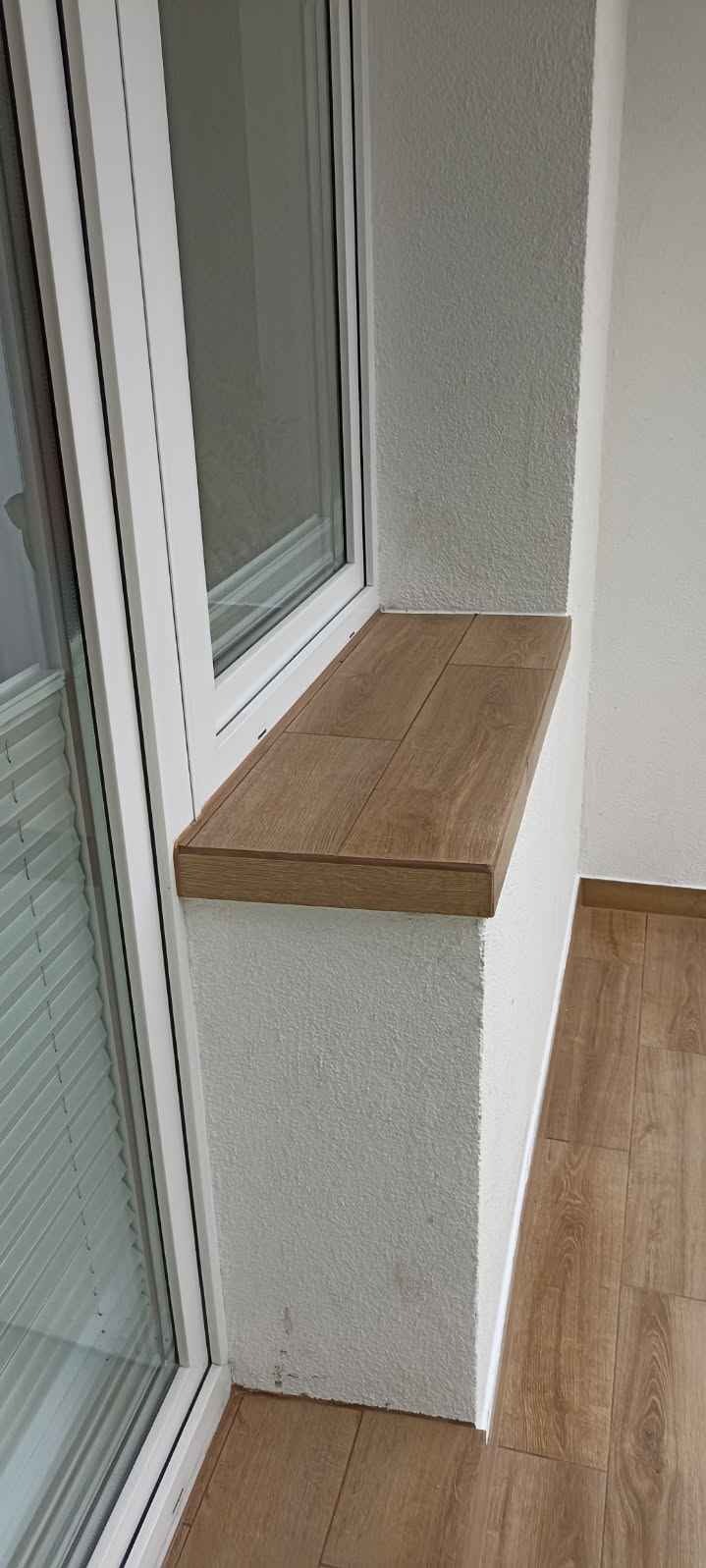 Wykończony parapet z płytek imitujących drewno na balkonie w Jabłonnie, dopasowany do podłogi z paneli. Widoczna biała rama okienna i struktura ściany.