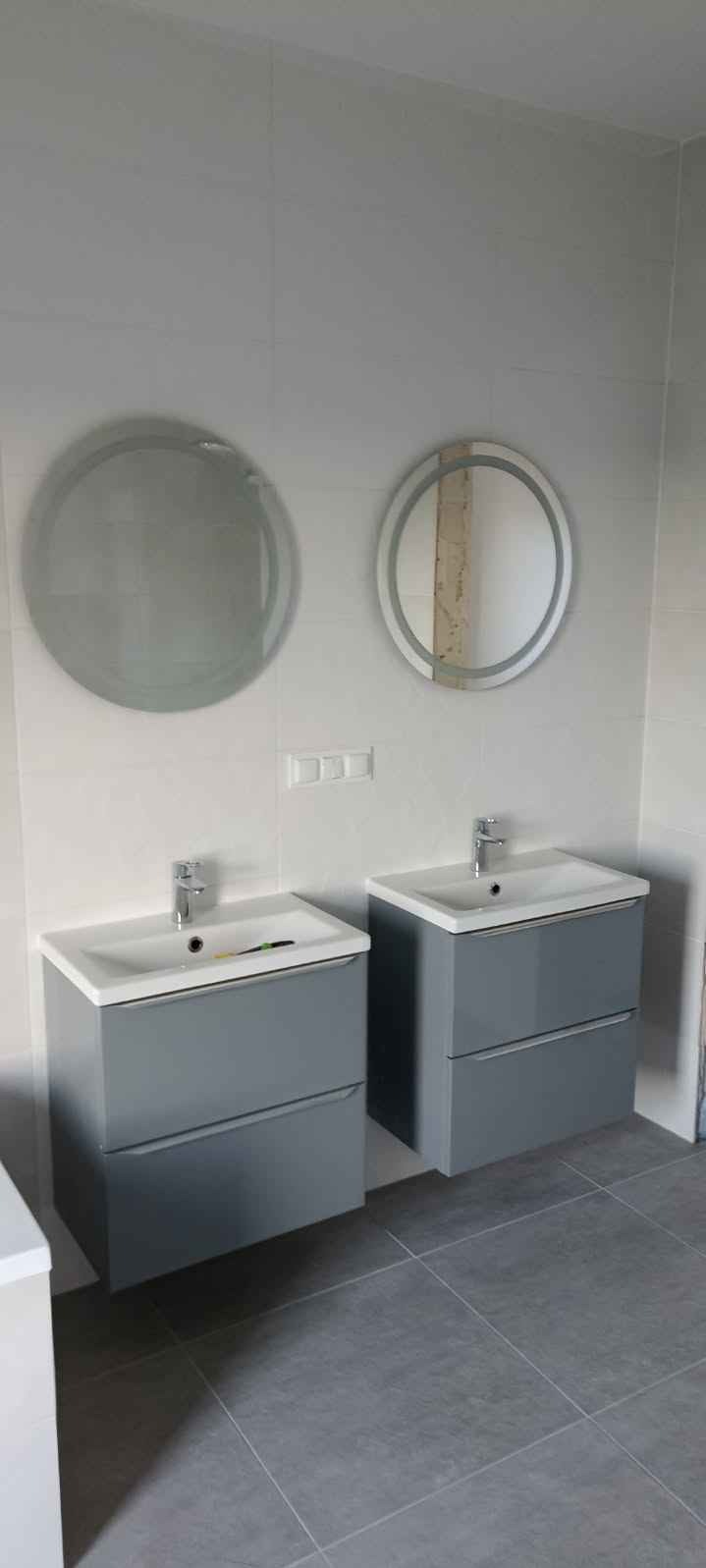 Dwie szare szafki łazienkowe z umywalkami i okrągłymi lustrami na białych płytkach, szare płytki podłogowe, minimalistyczny design.